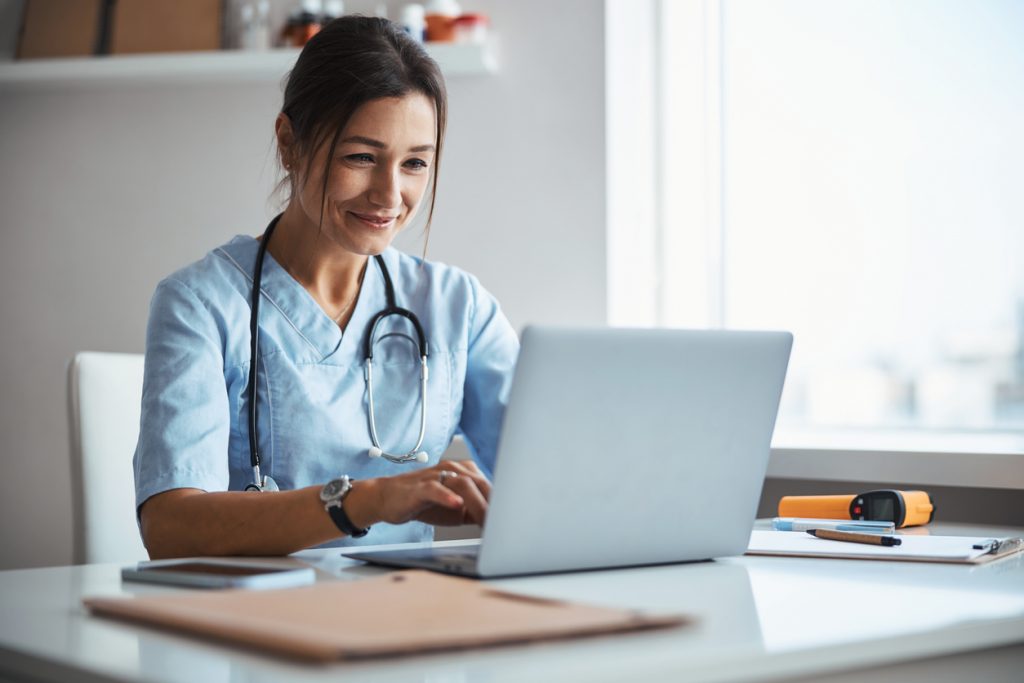 Beautiful young woman physician sitting at the table and smiling while working on modern laptop