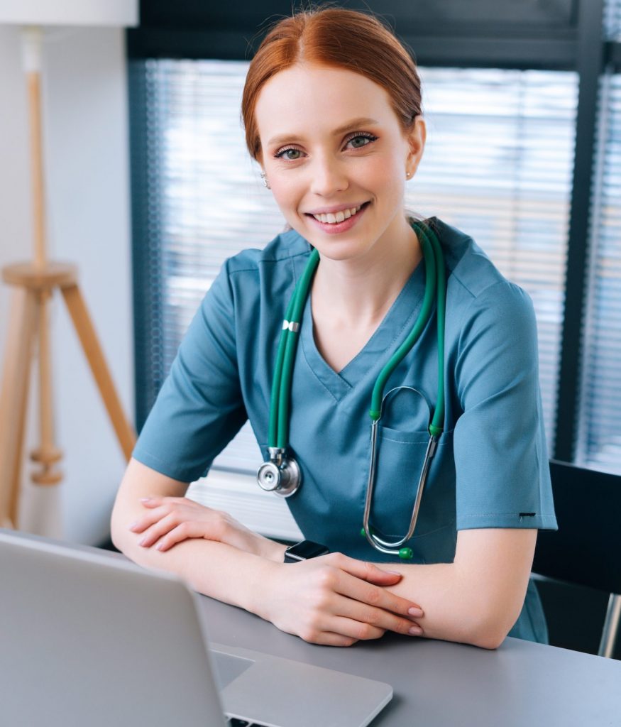 Close-up of red-haired smiling young woman in blue green medical uniform sitting at desk with laptop