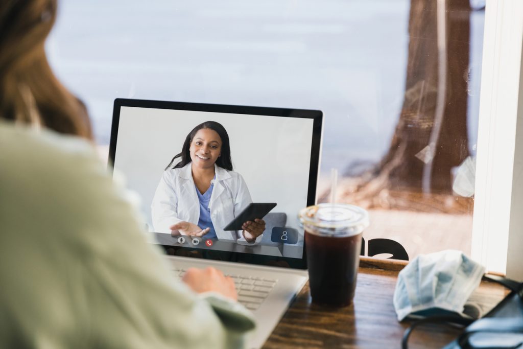 Because of the coronavirus outbreak, the unrecognizable woman uses a tele-medicine app on her laptop to consult with the mid adult female doctor.