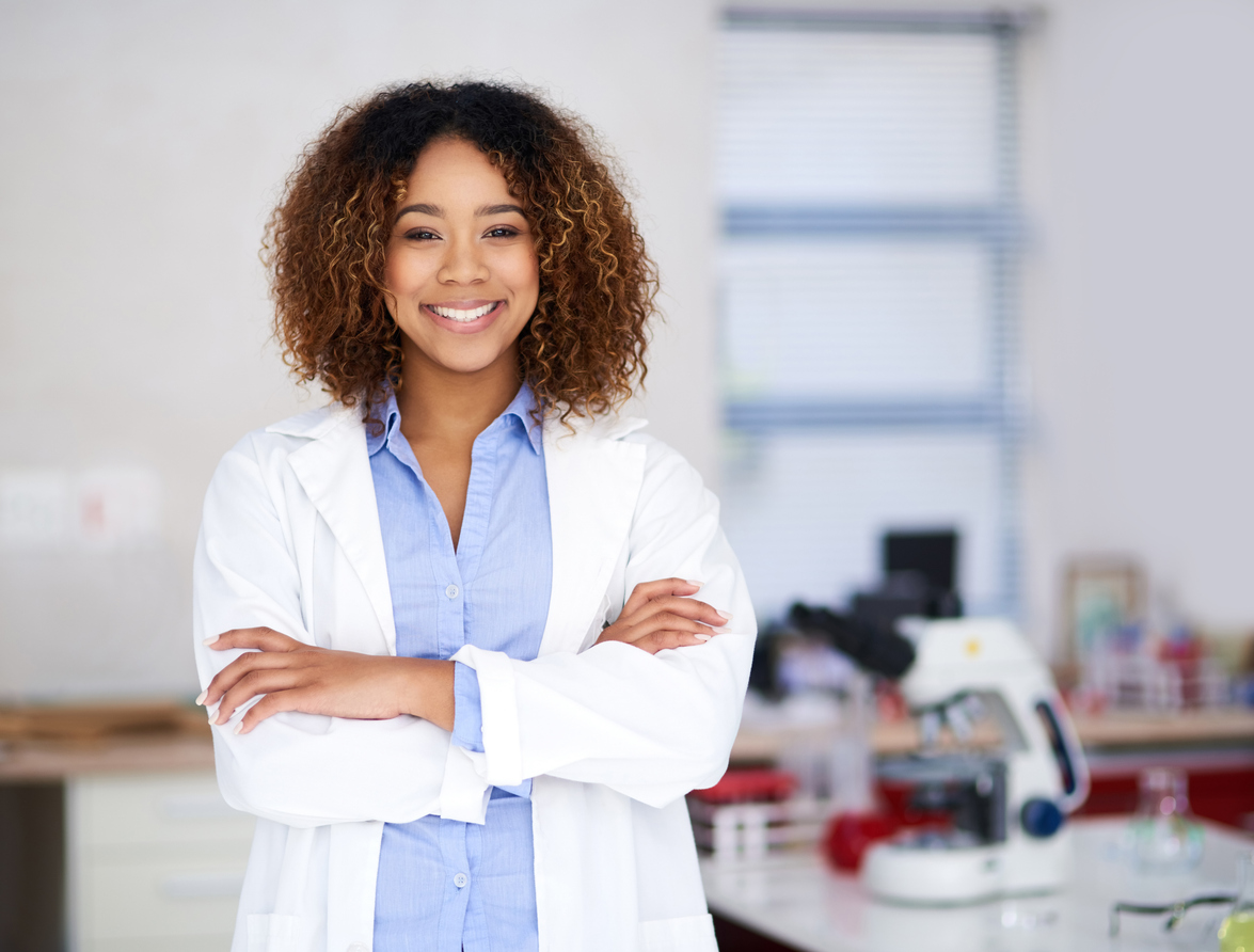 Woman in a white coat and blue shirt standing in a medical lab with folded arms